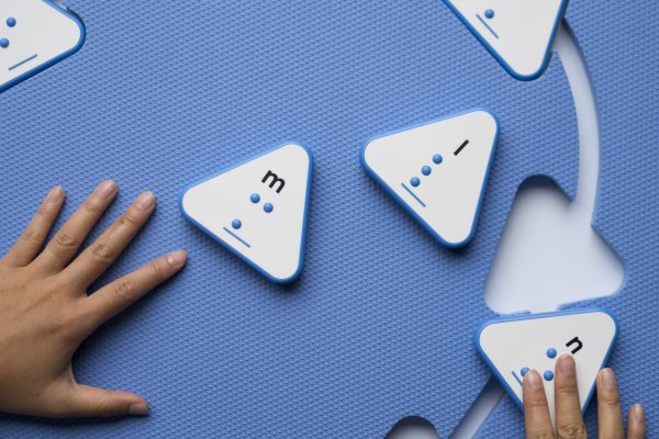 Overhead view of the blue mat with triangle cut-outs and a few blue triangular alphabet tiles. A child's hands are placing a tile into the cut out.