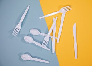 Three each of white plastic spoons, forks, and knives arranged on a yellow and light blue background.