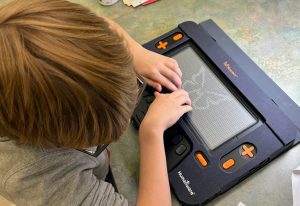 A young boy uses both of his hands to feel a tactile graphic of the butterfly featured in the Monarch logo on the Monarch’s 10 line by 32 cell refreshable braille display.