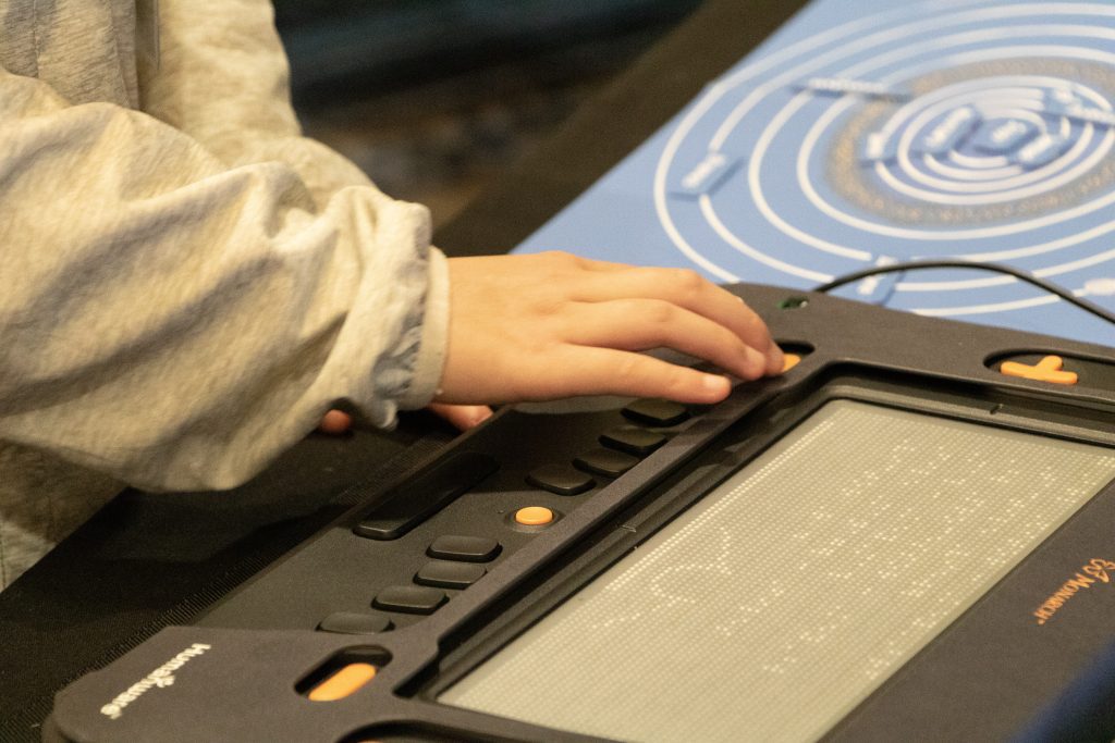 A student uses the Monarch's orange zoom out button while viewing a braille document. The top of a tactile graphic is visible at the bottom of the refreshable braille display. In the background a blue tactile solar system can be seen.