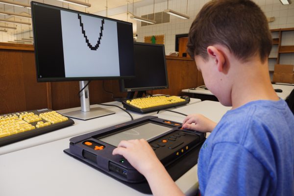 A young student in a library using a Monarch connected to a visual display. As he explores a tactile parabola graphed on the Monarch display using the KeyMath app, the same image is mirrored on a monitor above.