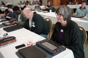 In a meeting room, people sit at long tables arranged in rows and covered in white tablecloths. Several of the people are examining the Monarch multiline braille displays in front of them.