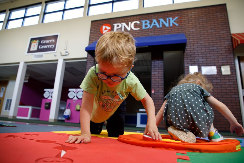 A young boy with glasses and another young child play on a Reach & Match mat. Behind them, a faux brick building facade with the PNC Bank logo at the top, mimicking a sign, can be seen.