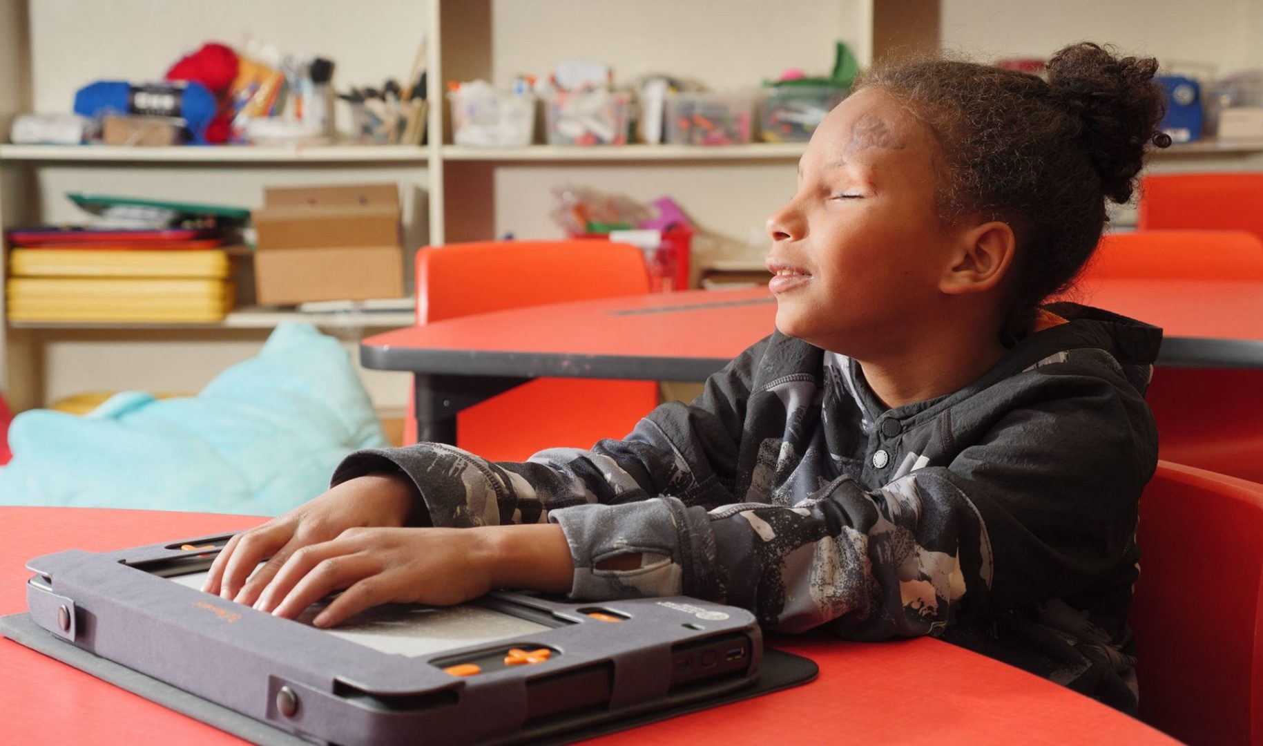 An elementary school-aged boy sitting at a table in a classroom looks pleased as he uses both hands to touch the Monarch’s multiline braille display.