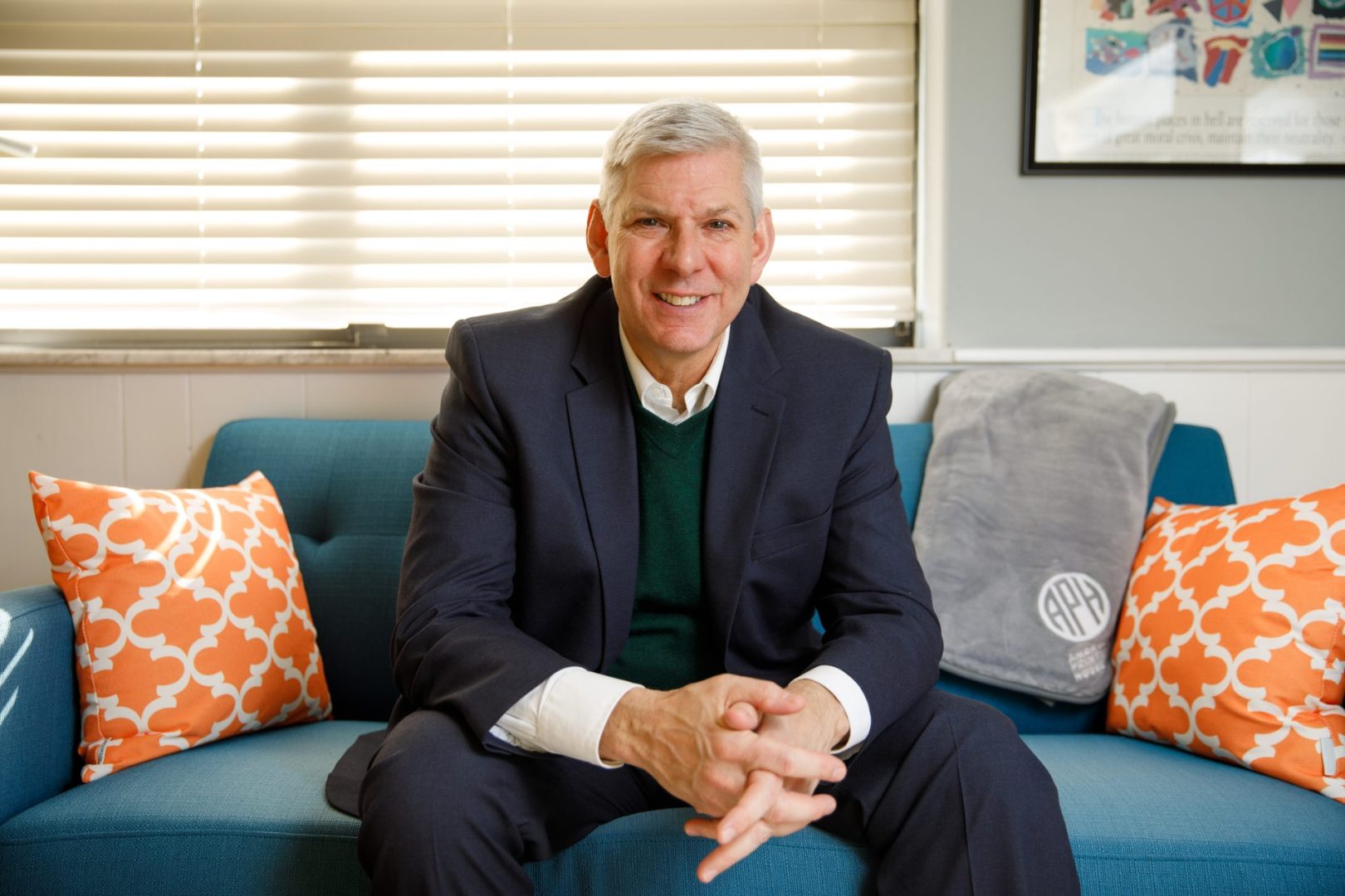 Craig Meador smiles as he leans forward to rest his elbows on his legs as he sits on a teal couch decorated with an APH branded blanket and orange throw pillows.