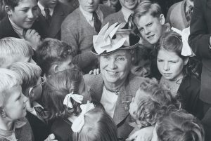 Helen Keller smiles while being surrounded by children in a black and white photo.