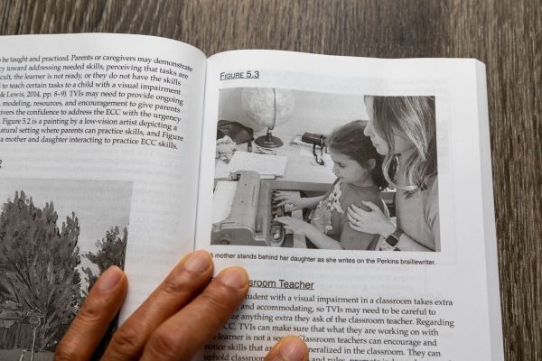 A hand holding open a copy of Tools for TVIs to a page showing a student working on a Perkins brailler with adult support.