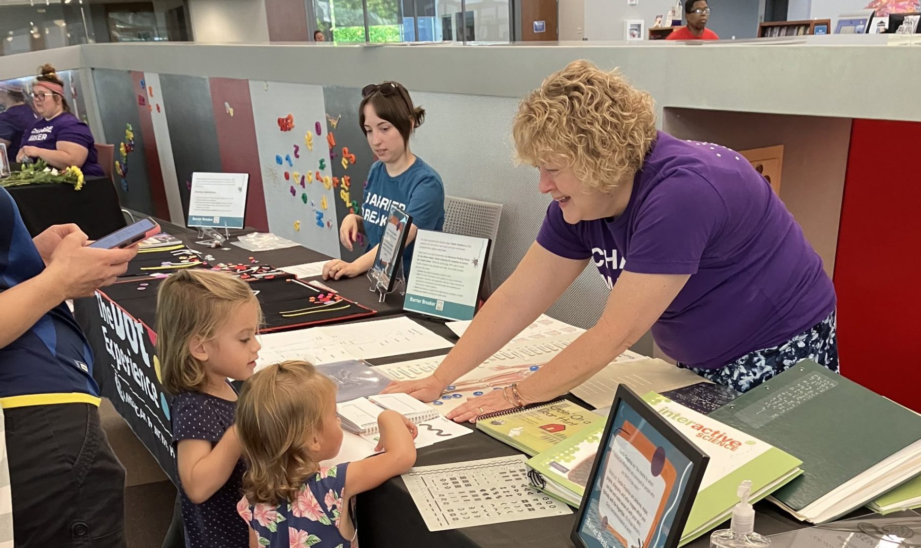 An APH employee leans over a table covered in papers and books as she speaks to two small girls and a woman.