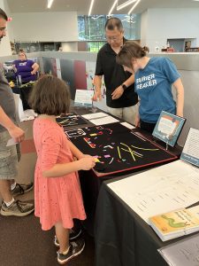 A young girl standing near a man plays with pieces of the  Picture Maker Wheatley Tactile Diagramming Kit, which are resting on a table, while two APH volunteers speak to her and the man from behind the table.