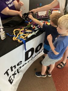 Two young boys stand with an adult in front of a table. The boy closest to the table and the adult reach for some textured rope being handed to him by an APH employee behind the table.