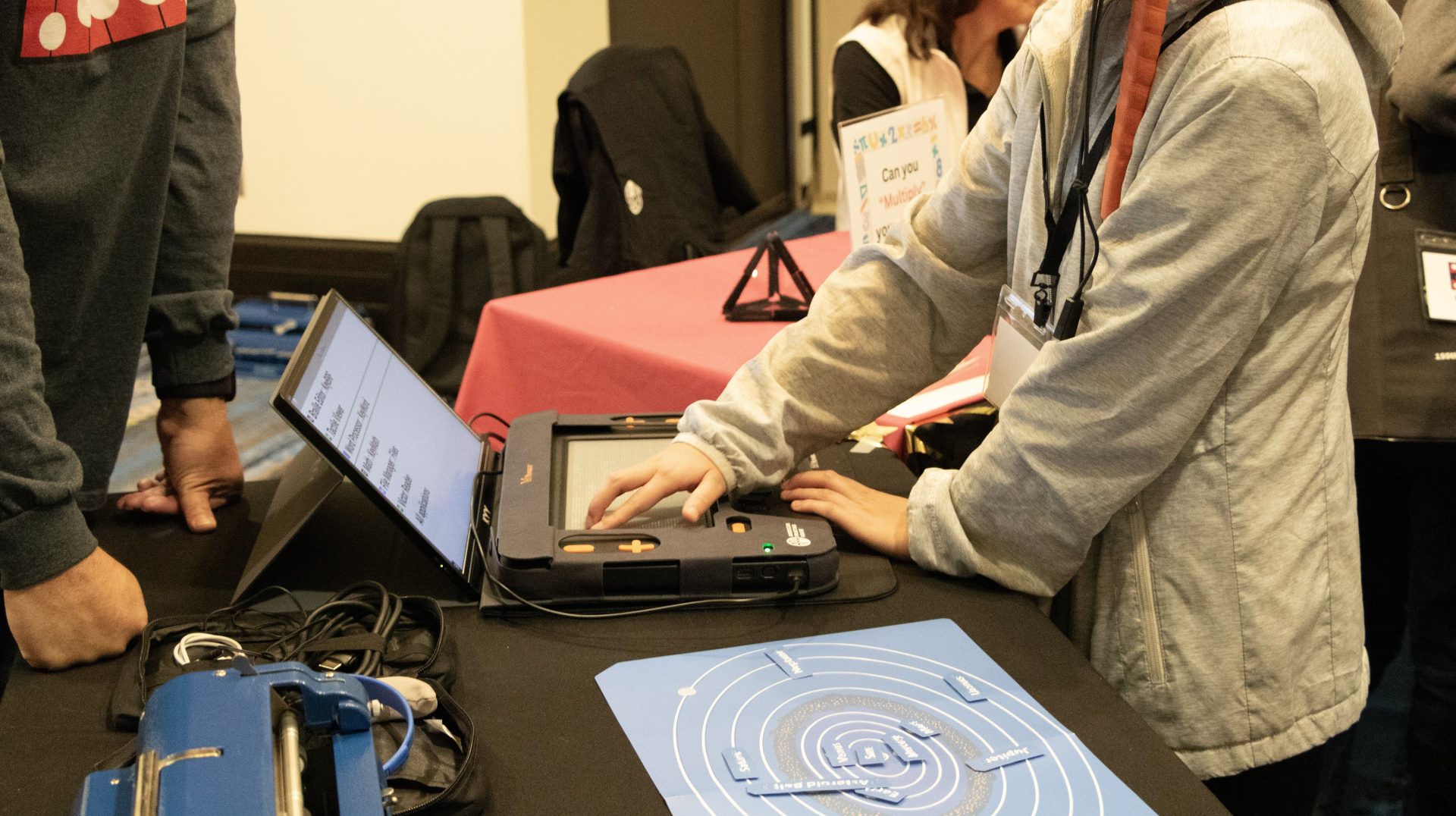 A middle school aged student with a white cane tucked into their arm touches the Monarch's multiline refreshable braille display, which is laying on a table in front of them. An adult stands behind the table, and a tactile solar system map and a Perkins brailler are also laying on the table.