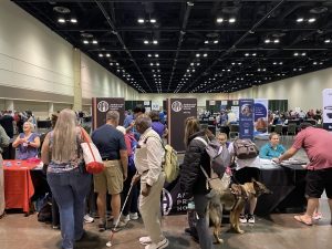 A large group of people, including a person with a guide dog, stand in front of the APH booth in a large convention exhibit hall.