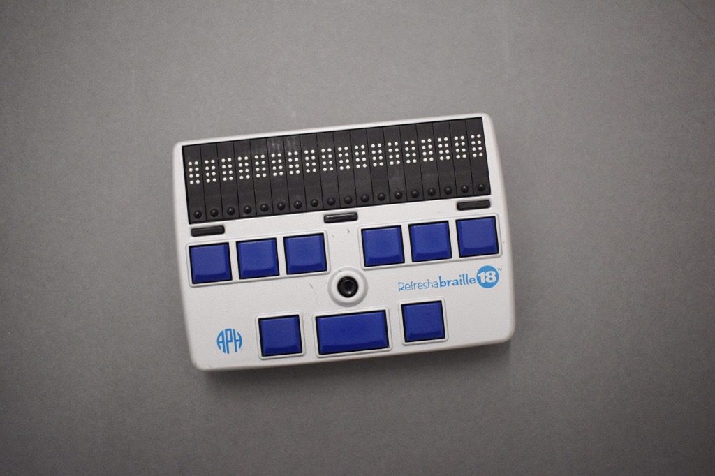 The Refreshabraille 18. The rectangular plastic case is silver and black with slightly rounded sides. An eighteen-cell, eight-dot refreshable braille display is at the top of the device, with a round cursor routing button below each cell and three display advance bars below the display. Nine square blue braille entry keys are arranged six over three beneath the display advance bars and the joystick, which is located in middle of the device. The APH logo and "Refreshabraille 18” in blue are visible on the silver case.