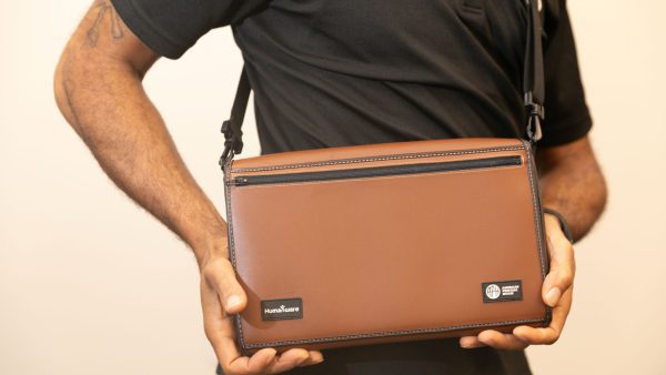 A man wearing a Mantis across his body in the Turtleback case and holding the closed device with both hands. The front of the brown case contains logos for HumanWare and APH.