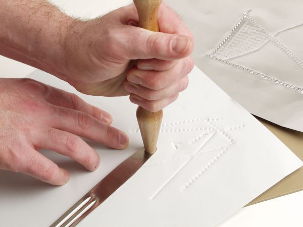 Two hands creating a tactile graphic of shapes and lines using the included hammer and tong tool on white paper.