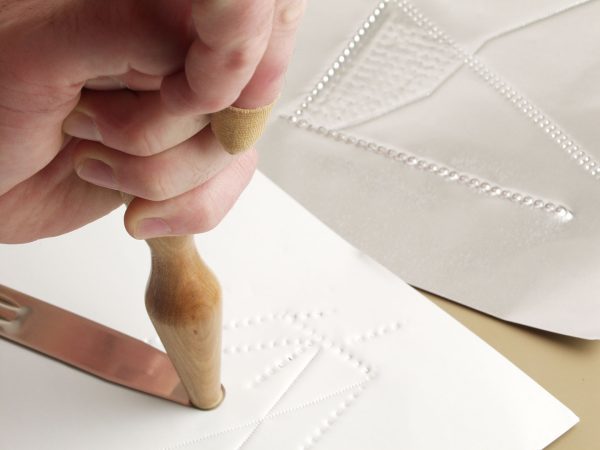 One hand holding the tactile graphics hammer against a point symbol on a white piece of paper.