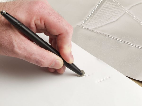 One hand creating a tactile line on a white piece of paper using the tactile graphics kit roller.