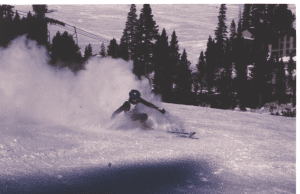 A skier makes a turn on a snowy mountainside with pine trees, whipping up snow behind him.