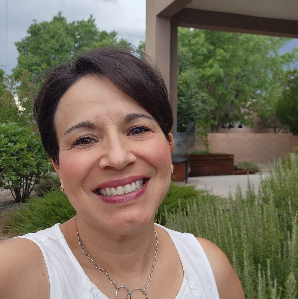 A dark haired woman with dark eyes wearing a white sleeveless top, a necklace, and earrings stands outside smiling at the camera.