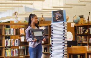 A student stands in a library next to a stack of braille notebooks with a Monarch in her hands.