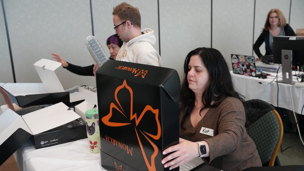 A young woman sits at a table with a Monarch box in front of her. She has lifted the lid on the box and is looking inside. A young man stands in the background.