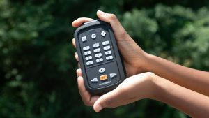 Two hands cradling an open VR Cardinal device against a background of dark green foliage. On the face of the device are various buttons, and along the bottom underneath the play button, “VictorReader” is printed in white.