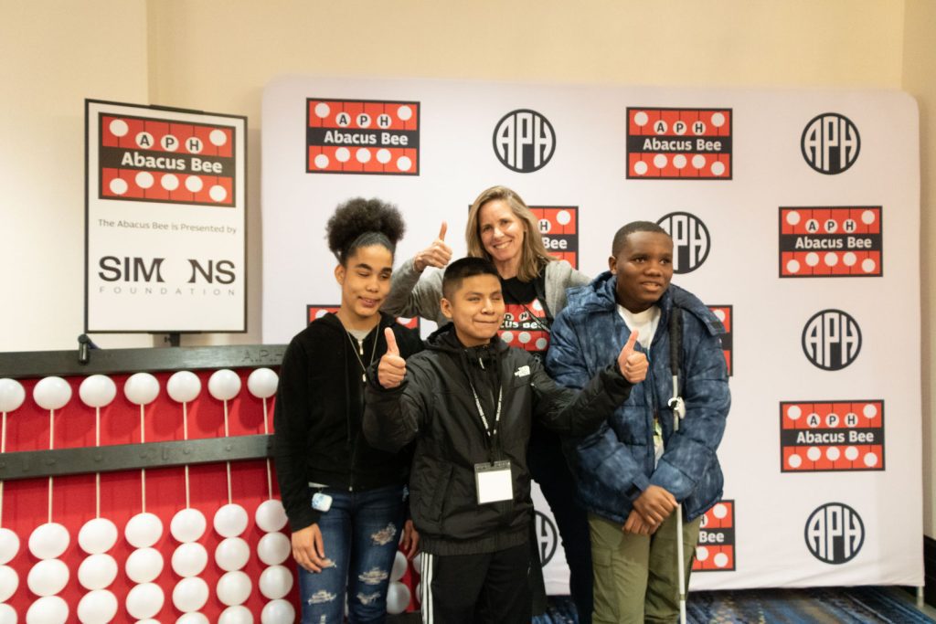 A group of three students and an adult stand together, smiling and giving thumbs up in front of a backdrop with the APH and Abacus Bee logos. Behind them to the left is a giant abacus and a sign with Abacus Bee and Simons Foundation logos