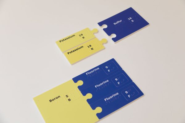 Two sets of bonded subunits. On top are two yellow potassium subunit pieces sitting next to a large sulfur piece. The element's name, atomic number, and atomic symbol are printed and embossed.