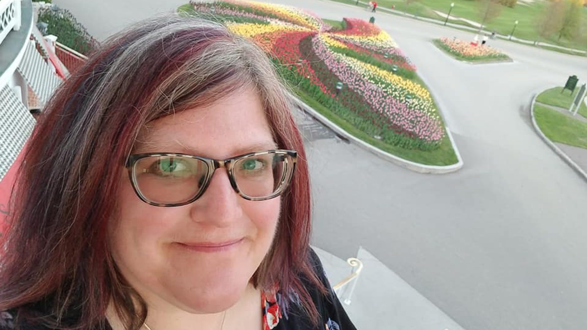 A young woman with red highlighted hair and glasses smiles at the camera. Flowers are in the background.