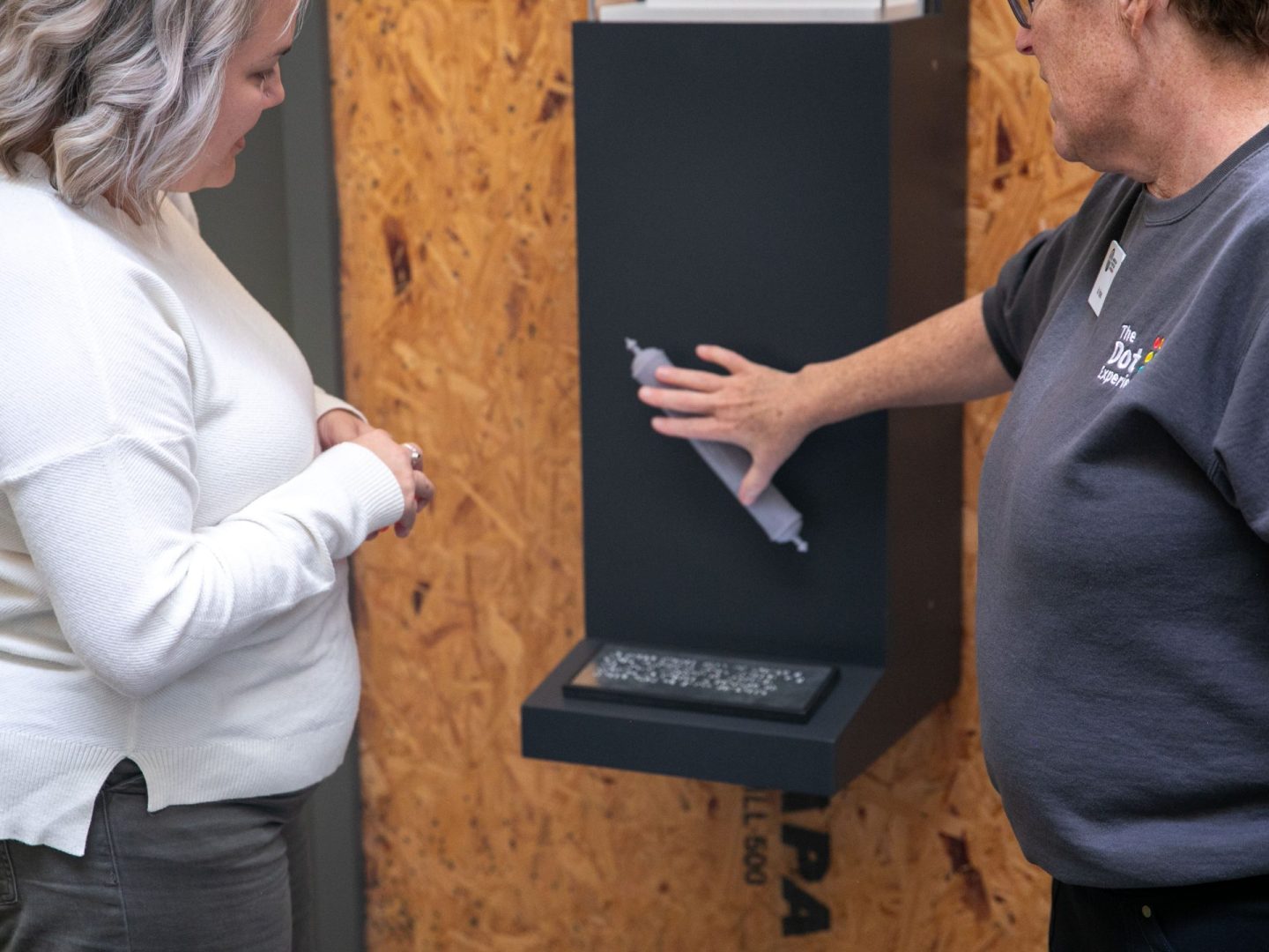 Two women stand in front of the scroll cast as one feels the tactile graphic.