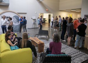 Guests stand in a room with exhibits, a yellow couch, and many chairs as they look around the room.