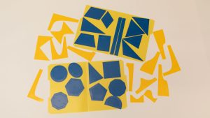 An overhead shot of the Geometric Drawing Stencils attached to a yellow folder with the 3D printed function stencils spread around them. Each shape has an inner stencil and an outer stencil. Shapes include ovals, octagons, circles, triangles, and many more polygons.