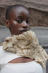 A woman stands, wearing a white shirt with a textured piece around her neck in a shape similar to a seashell.