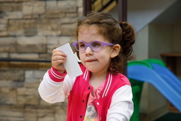 A preschool girl in a white and pink letterman-style jacket and purple glasses holds a crescent shape puzzle piece up to her face to look at it.