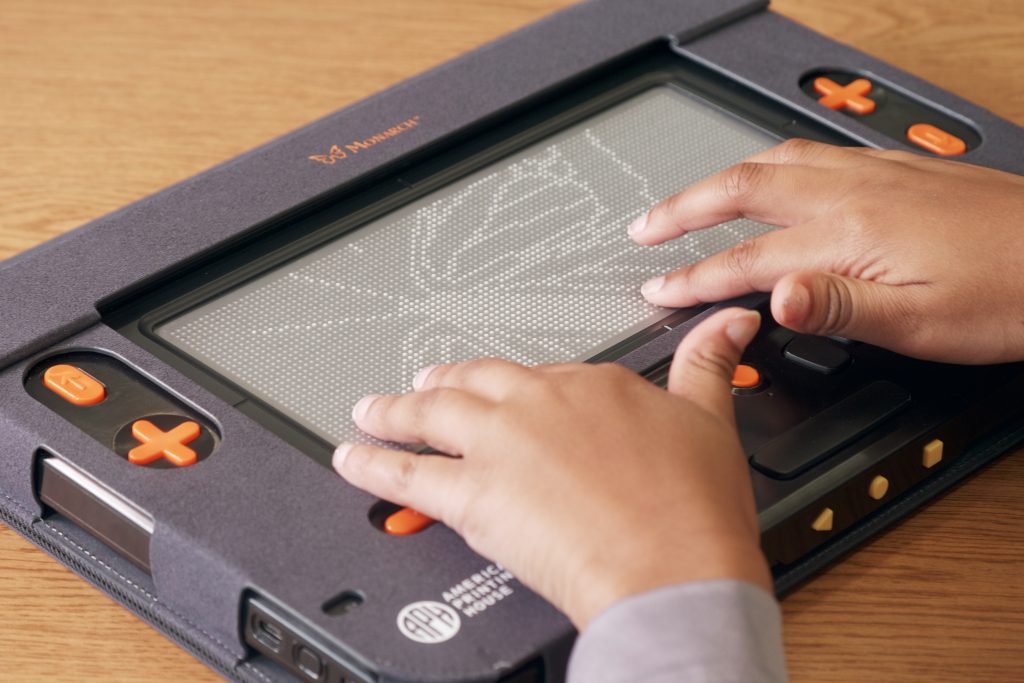 Two hands rest on the Monarch, feeling the lines of the refreshable braille tactile butterfly graphic.