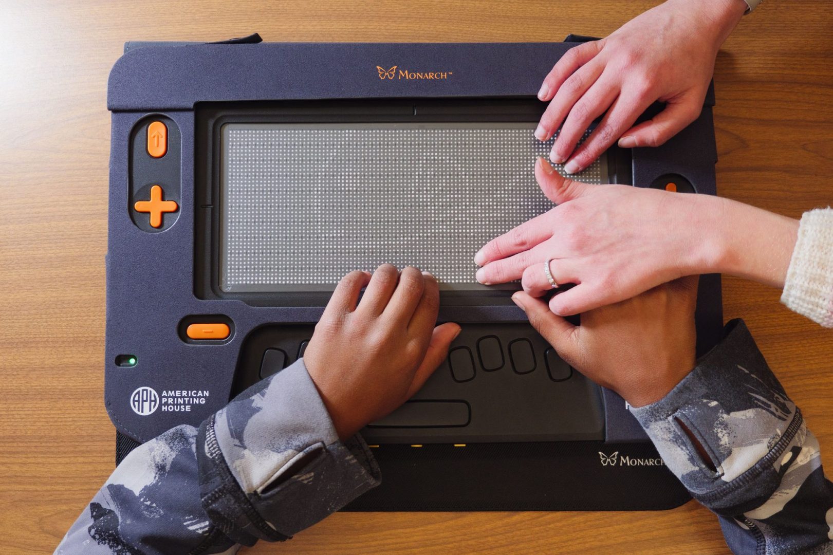 A young student in a black and grey hoodie has two hands on a Monarch device sitting on a wooden table. An adult woman is guiding his right hand across the tactile surface. Her other hand rests on the top left corner of the Monarch.