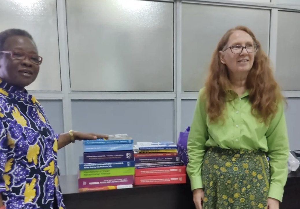 Two women, one of which is Dr. Elyse Connors, smile as they stand near two short stacks of book resting on a table.