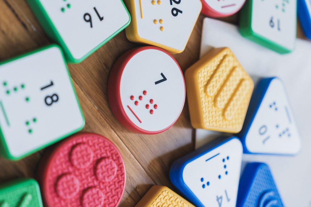 An assortment of Reach and Match number tiles spread out across a wooden floor. In the center is a red circular tile with the number 