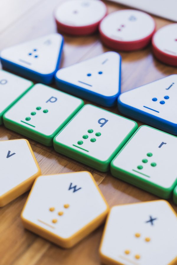 Four rows of Reach & Match Alphabet Tiles on a wooden floor. The row of square green tiles are in focus with blue triangles and red circles above and a row of yellow hexagonal tiles beneath.