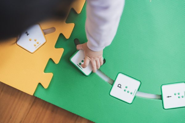 A child's hand inserts a green square alphabet tile into the green mat on a wooden floor.