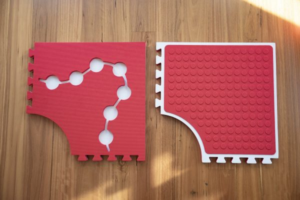 Two red Reach & Match mats side by side demonstrating the differences between the textured side and the side with circular cut-outs for tiles and a traceable path.