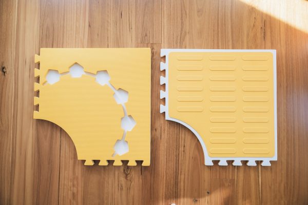 Two yellow Reach & Match mats side by side demonstrating the differences between the textured side and the side with hexagonal cut-outs for tiles and a traceable path.