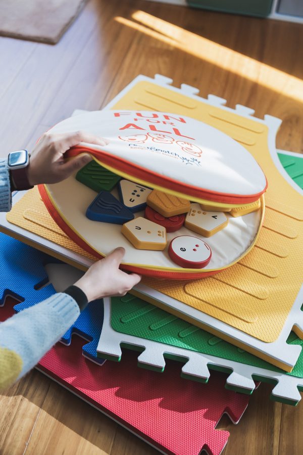 On top of four stacked Reach & Match Light tiles on a wooden floor, two hands open the zip carrying case that contains the Reach & Match Alphabet Tiles.