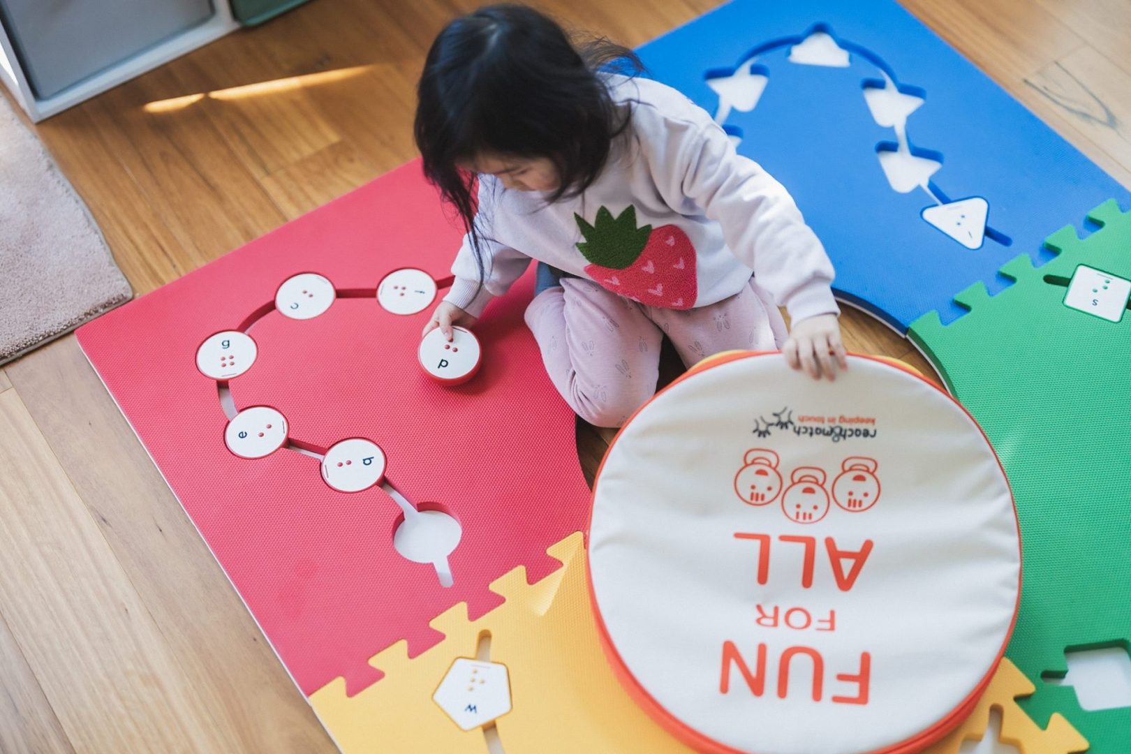 Photo from above of a young girl sitting on the reach and match tiles with a circle 'd' in her right hand and holding the reach and match case in her left hand.