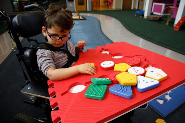 A little boy in a wheelchair with a red Reach and Match foam square on top of his wheelchair tray with a variety of tiles.