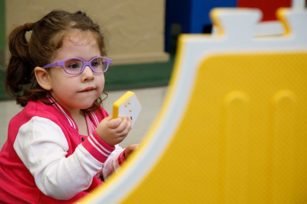 A little girl in purple glasses holding a yellow Reach and Match tile up to her eyes while making an inquisitive facial expression.