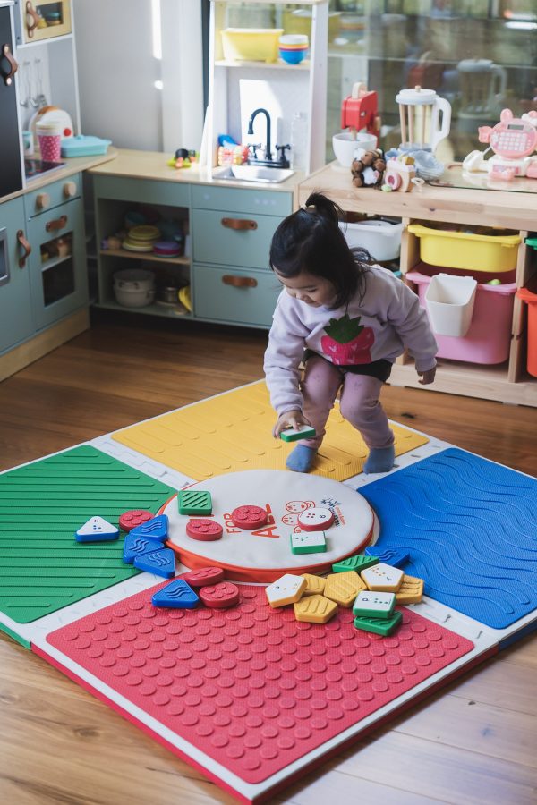 A little girl plays with Reach and Match tiles assembled in a square with their textured sides visible. The tile carrying case is placed in the center of the mats.