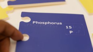 Close up of the blue Nemeth Phosphorus piece. The name of the element is printed across the top in print and braille, and there is a raised box in the center right of the piece that contains the atomic number and the atomic symbol.