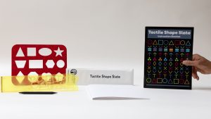 All the components of the Tactile Shape Slate kit, including a yellow slate, a large red stencil, and the guidebook, all laid out on a white background.