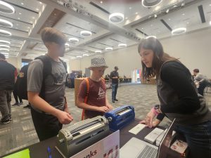 A young woman stands behind a booth while two boys watch her explain the products on the table.